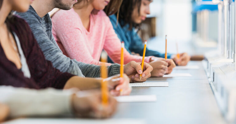 Four teacher candidates are seated at a long desk. Each has a pencil, which they are using to take the Praxis exam.